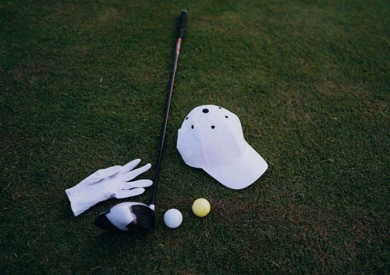 two golf balls, golf glove, white cap and golf club laying on the grass