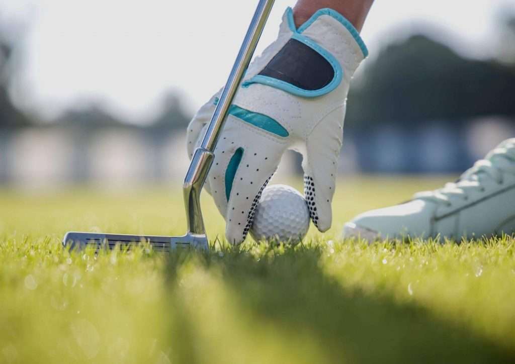 a hand with a golf glove picking up a golf ball on the grass while holding a golf club